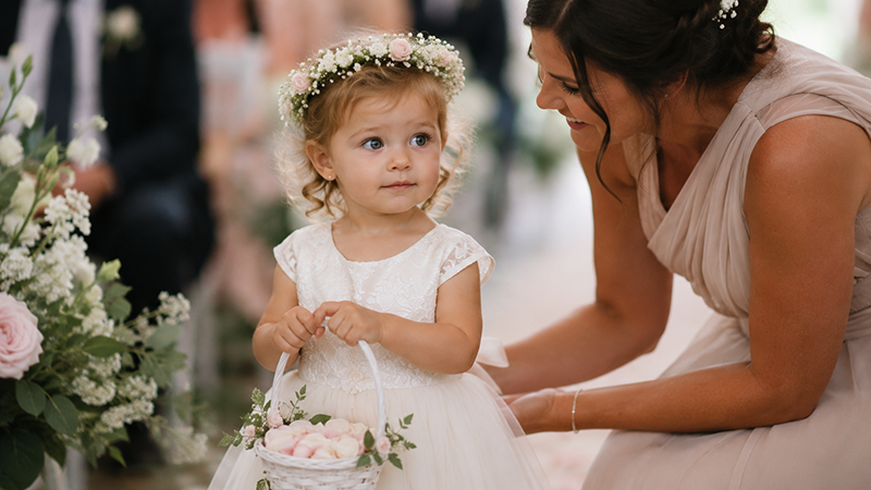Two-year-old flower girl holding a basket while standing beside her mother before a wedding ceremony