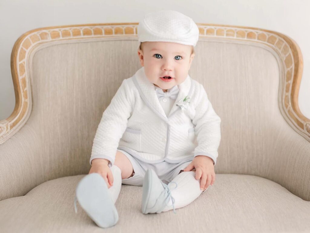 Baby boy wearing a white baptism outfit with a hat, sitting on a chair